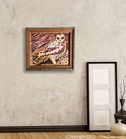 Original oil painting of an owl hung on a textured wall next to a framed set of blank artworks and a vase with branches.