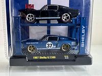 Front view of unopened package showing both black and blue 1967 Ford Shelby GT500 diecast cars on a blue auto lift display stand.