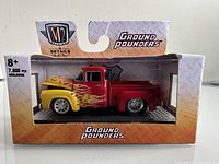 Front and side view of unopened M2 1956 Ford F-100 Truck diecast model in original packaging with yellow front and red rear with flames