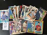 Photo showing a collection of 27 assorted baseball cards laid out on a dark surface, including images of various players such as Gary Carter, Joe Rudi, Gary Sheffield, and others.
