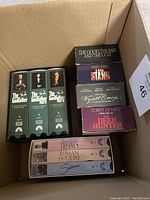 Six VHS movie sets in a cardboard box including The Godfather Trilogy, The Good The Bad and The Ugly, Stephen King's IT, Wyatt Earp, The Deer Hunter, and a three-movie set of Tiffany's, Roman Holiday, Sabrina.