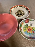 Three bowls placed on carpet. One large pink plastic bowl, one medium floral printed plastic bowl, and one white ceramic bowl with vintage Pasco logo with red and green rim stripes.