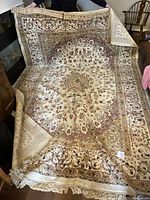 Large rectangular Persian Tabriz area rug displayed atop table, showing beige field with central medallion and floral borders, fringed ends