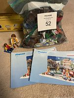 Playmobil Christmas figures including Santa, child on sled, adult figures, and two instruction booklets on floor with bag of mixed figures and accessories.