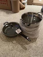 Large stainless steel stock pot with glass lid next to a large covered non-stick skillet shown on carpeted floor.