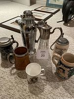 Photo showing varied drinkware including the crystal and silver carafe, ceramic beer stein, wooden tankard mug, and several mugs including the Queen Elizabeth II tea cup.