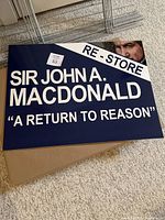 Close-up image of a Sir John A. MacDonald lawn sign showing text and partial face image on navy blue background.