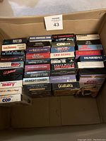 Photo shows 35 assorted VHS movie tapes stacked inside a box, titles visible on spines and covers.