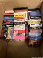 Photo showing a full box with 34 VHS movie tapes arranged both vertically and horizontally, titles clearly visible on most tapes including Golden Eye, Raw Deal, The Full Monty, Wayne's World, Liar Liar, and others.