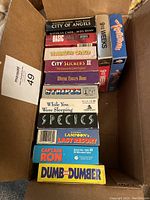 Photo of 13 VHS movie tapes stacked inside a cardboard box. Titles visible include Dumb and Dumber, Basic Instinct, Species, While You Were Sleeping, Stripes, City Slickers II, City of Angels, National Lampoon's Last Resort, Captain Ron, and 9 1/2 Weeks.