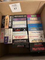 Photo showing a box containing 25 VHS movie tapes neatly arranged with visible spines labeled with various movie titles.