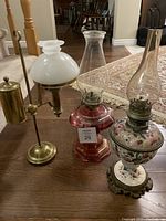Three vintage oil lamps displayed on a wooden floor: cranberry glass lamp, floral porcelain lamp, and brass adjustable lamp with white globe.