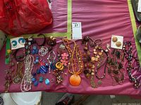 Wide shot of assorted costume jewelry spread on a pink and yellow mat, showing necklaces, earrings, bracelets in various styles and colors including orange, blue, brown, and beige.