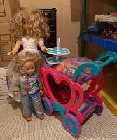 Both dolls, pink and blue plastic tea trolley loaded with dishware and clothing in clear bag