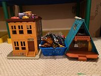 Full lot overview showing the Fisher Price Sesame Street building, blue plastic basket with toys including a brown horse and assorted figures, and a separate blue A-frame plastic building.