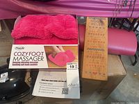 FineLife Cozy Foot Massager boxed with visible product and packaging including bright pink plush foot massager with bow, next to wooden laptop desk with printed house rules and rolled pink Lululemon yoga mat behind.