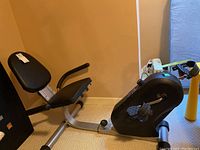 Side view of black and silver recumbent exercise bicycle showing padded seat and backrest, resistance knob, and pedals with straps.