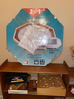 Full view of boxed deluxe Crokinole game with visible packaging and some unpackaged games below on wooden shelf including backgammon and wooden maze game.