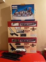 Stacked boxes showing Lemax Village Train sets with circular tracks, Lemax skating pond box on top, and extra black plastic train tracks laid out in front