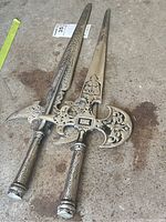 Two steel halberd polearm end replicas laid on ground showing overall shape, size and intricate engraving.