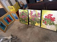 Three floral paintings and rock and roll triptych with hinged frames arranged on floor in garage
