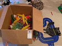 Box containing various plastic toy food items including bananas, grapes, corn, and other vegetables, along with a plastic toy race track partially visible next to the box.
