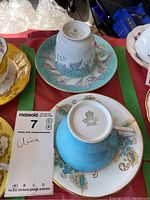 Photo shows two teacups and saucers on a red surface, one turquoise with grey birds and flowers (Wedgwood), the other turquoise and white with gold floral accents (Ainsley), photographed with focus on markings and design details