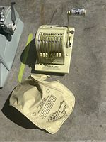 Top view of the Paymaster cash adding machine with visible number dials, hand crank, and dollar/cent keys, alongside its branded cloth cover placed beside it.
