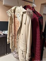 Three women's coats hanging on a hanger including beige leather parka, plum quilted jacket, and black red plaid coat.