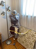 Dressmaking model with plastic cover, adjustable metal stand, and instructions attached, positioned next to the floor lamp and folding table with floral cushion in a room with wooden floor and curtained window.