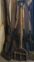 Photo showing multiple garden tools including shovels, garden forks, hand picks, and a garden seeder stored in basement corner.