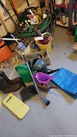 Wide view showing green garden hoses coiled, watering cans, black boots, yellow buckets with brushes and cloths, black scoop, yellow kneeling pad, blue/orange tarp, gray crate, and garden hand tools spread around