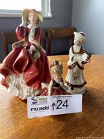 Three porcelain figures on a wooden table, including a large Royal Doulton lady in red dress, medium-sized lady with harp and small child figure in dress.