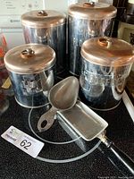Four vintage metal caddies labeled Flour, Sugar, Coffee, and Tea, arranged on a kitchen surface with two metal scoops in the foreground. The caddies have copper-colored lids and display wear and tarnish consistent with age.