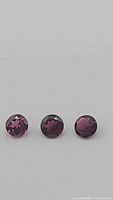 Close up photo of three round brilliant cut rhodolite garnet gemstones with deep electric purple color on white background.