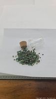 Small untreated emerald crystals in various small rough forms spilled next to a small clear glass jar with cork stopper on white background.