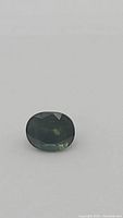 Close-up image of an oval faceted green sapphire with blue undertones on a plain white background showing stone details and color.