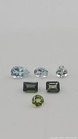 Close-up of six faceted gemstones: three aquamarines (pear, oval, round), two green rectangular tourmalines, one round peridot