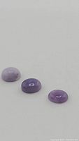 Three natural purple opal gemstones arranged on a plain surface showing their oval cabochon cut with variation in purple color tones and translucency.