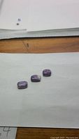 Three cushion cabochon purple opal stones placed on a white sheet atop a wooden surface, showing overall size and coloring of stones.