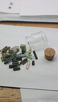 Loose assorted natural terminated tourmaline crystals on white background next to glass vial and cork stopper.