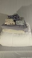 View of stacked bedding items showing white queen-sized duvet on bottom, beige and gray flat sheets folded on top with two patterned pillowcases folded atop.