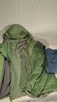 Photo showing green Columbia coat with hood, unzipped to reveal inner shell and part of a folded pair of blue pants.