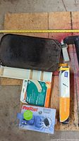 Overview of the lot showing black griddle, white easel frame, boxed remote car starter kit, small phone holder accessories, and small black pump parts on plywood surface.