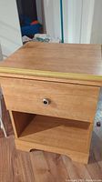 Full view of pressed wood nightstand showing drawer and open shelf, top surface with some wear and gold trim detail.