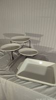 Photo of three-tiered serving tray stand with round white ceramic tiers on metal legs and two white rectangular trays in front