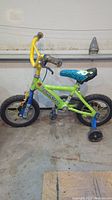 Full side view of toddler bike with green frame, yellow handlebars, blue fork, and blue training wheels. Tires appear deflated.