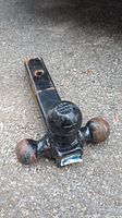 Black Reese Tri-Ball trailer hitch resting on gravel ground, showing one side with three tow balls of differing sizes, surface rust and wear marks visible.