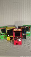 Five handheld arcade style video games standing upright, showing front control faces and screen graphics for each game: Frogger, Pac-Man, Mortal Kombat, Defender, Galaga.