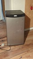 Full view of stainless steel Whirlpool mini fridge with black top edge, standing on wooden floor.